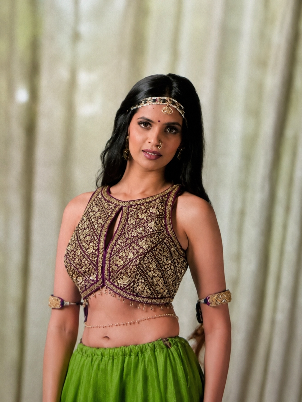 A woman modeling a traditional Indian outfit featuring a heavily embroidered brown and gold sleeveless crop top with a keyhole cutout, paired with a bright green skirt. She is accessorized with a gold headband, bangles, and a waist chain, set against a soft beige curtain background.
