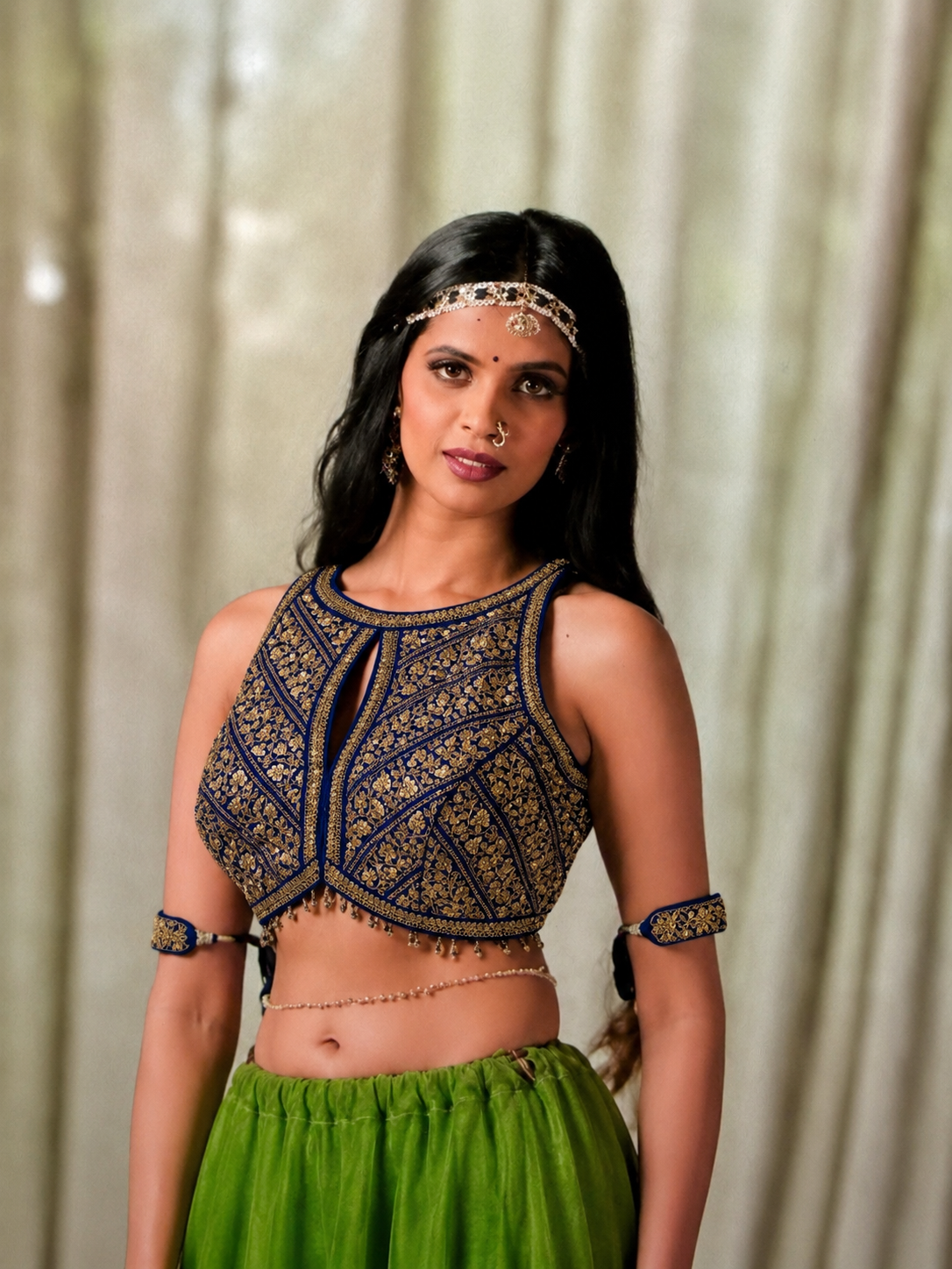 A woman modeling a sleeveless dark blue and gold heavily embroidered crop top with a keyhole cutout, paired with a green skirt, set against a soft beige curtain background.