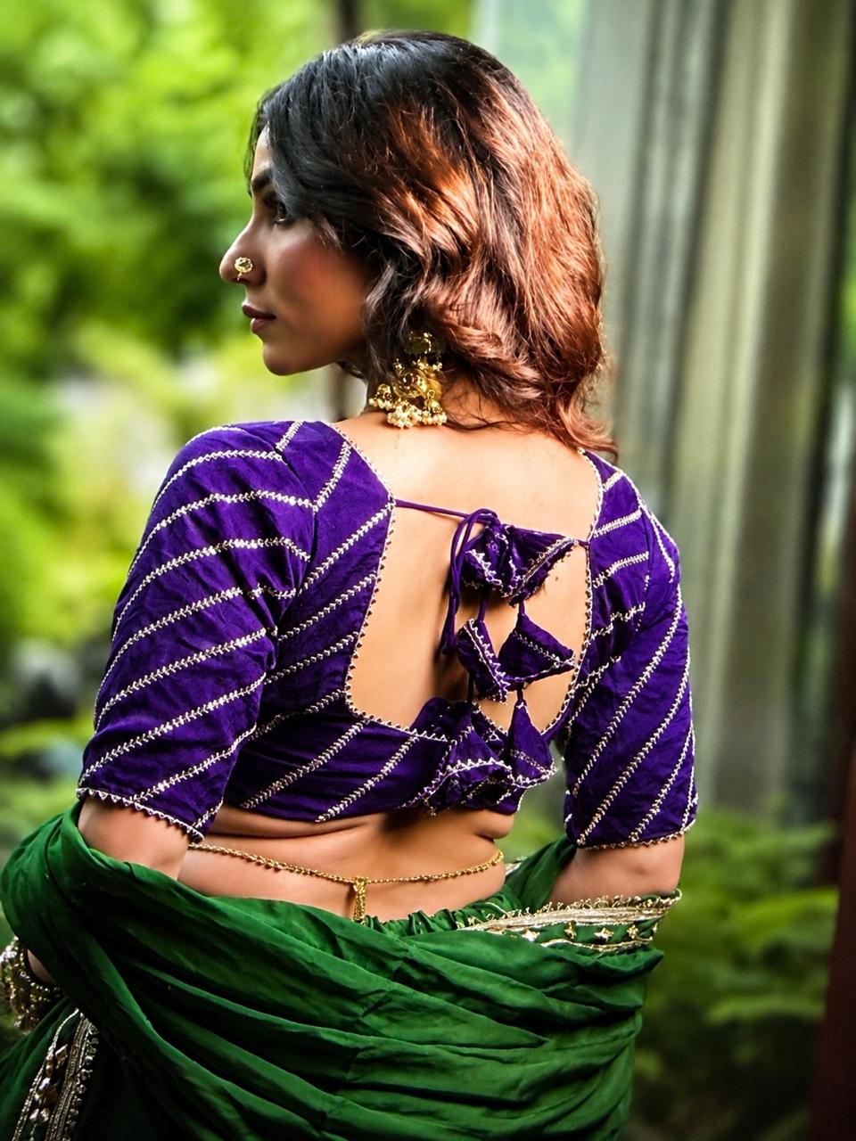 A woman shown from the back wearing a purple velvet blouse with silver stripes and a green sari, set against a blurred green outdoor background.