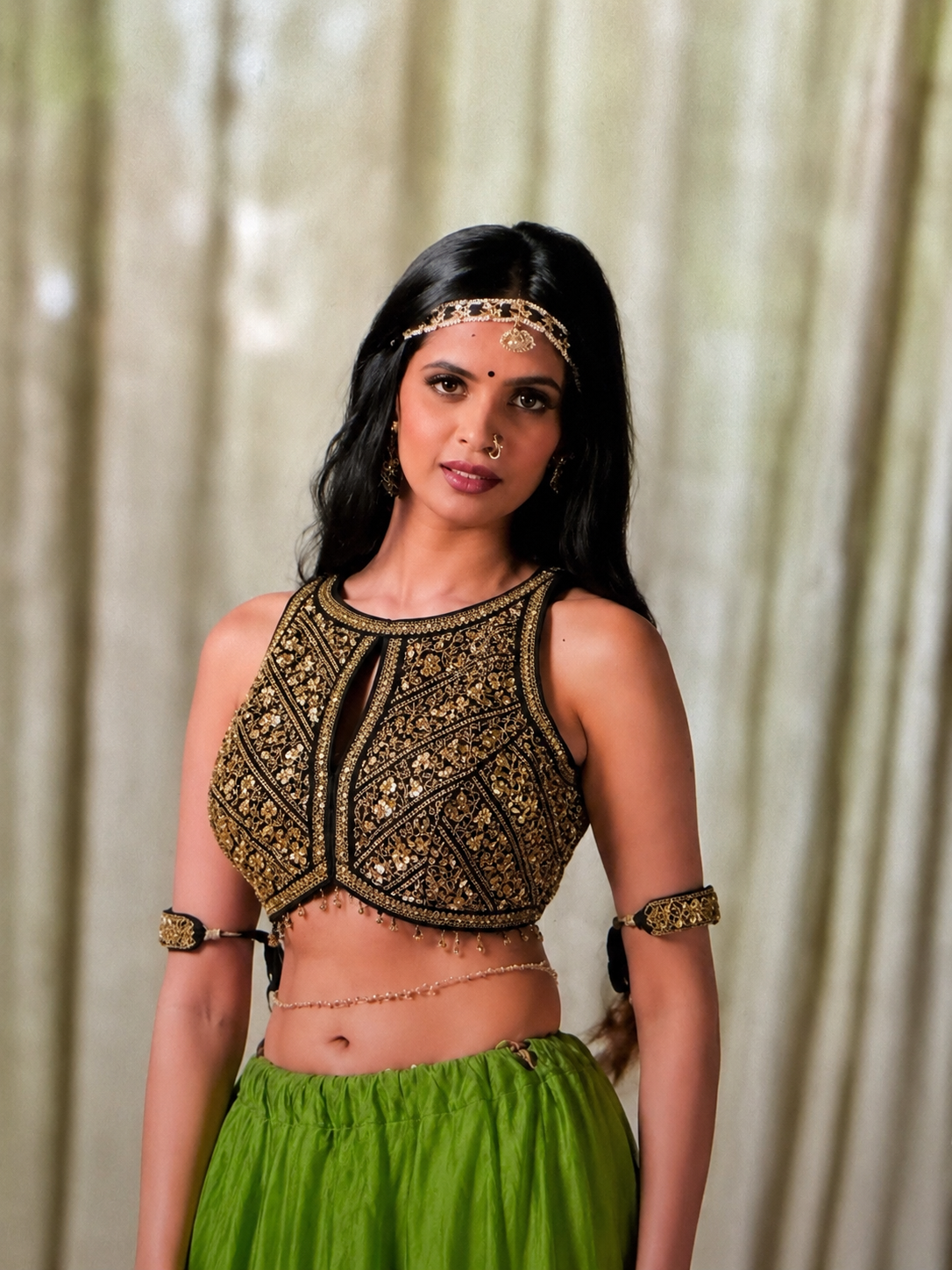 A woman modeling a black and gold heavily embellished sleeveless crop top with a keyhole cutout, paired with a green skirt, set against a soft beige curtain background.