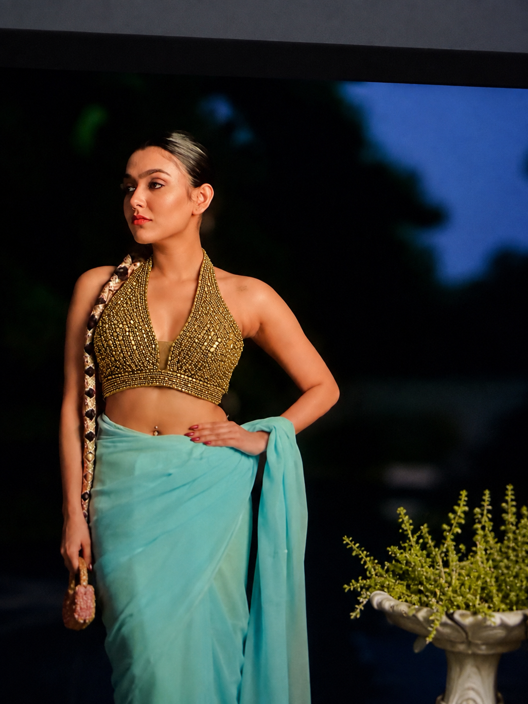 A woman modeling a light blue saree with a heavily embellished gold sequined halter-style top, standing against a dark background with a decorative plant pot at the side.