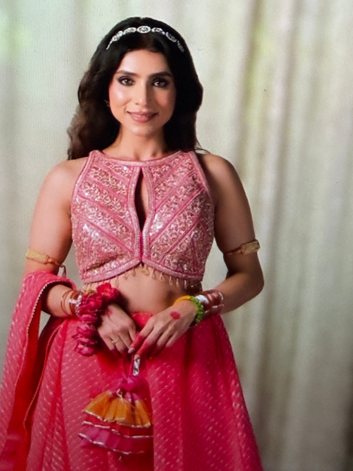 A woman modeling a traditional pink lehenga choli outfit with intricate gold embroidery, a matching pink dupatta, and colorful bangles, set against a soft beige curtain background.
