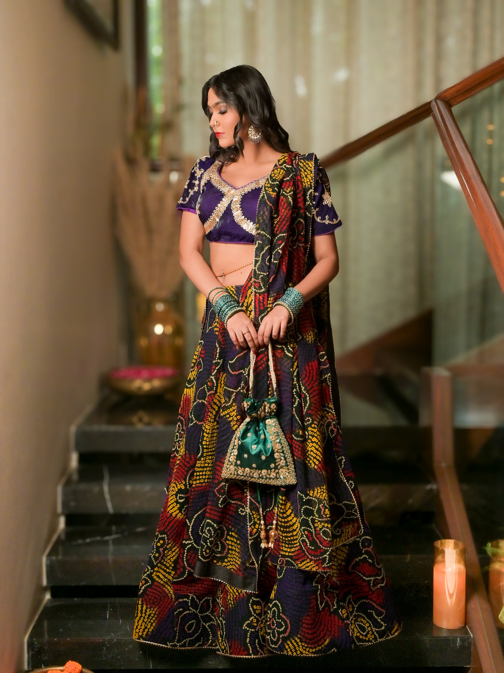 A woman stands on indoor stairs wearing a heavily embroidered traditional outfit with a purple blouse and a long multicolored printed skirt and dupatta. She is holding a small embellished pouch, with warm ambient lights and a staircase railing in the background.