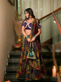 A woman stands on indoor stairs wearing a heavily embroidered traditional outfit with a purple blouse and a long multicolored printed skirt and dupatta. She is holding a small embellished pouch, with warm ambient lights and a staircase railing in the background.