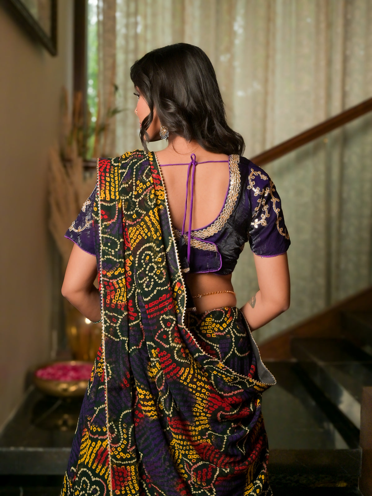 A woman shown from behind wearing a purple and black patterned saree with a matching embroidered blouse in an indoor setting.
