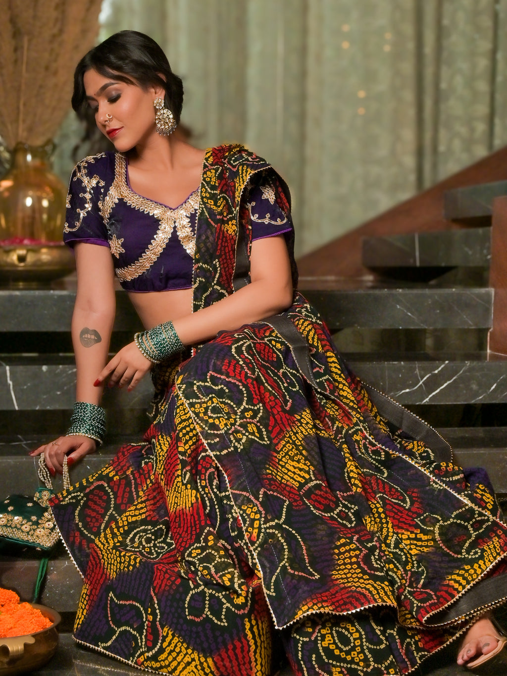 A woman models a traditional Indian ethnic outfit consisting of a purple embroidered blouse and a highly patterned flared skirt in purple, yellow, red, and black, styled with green bangles and earrings while sitting on stone steps.
