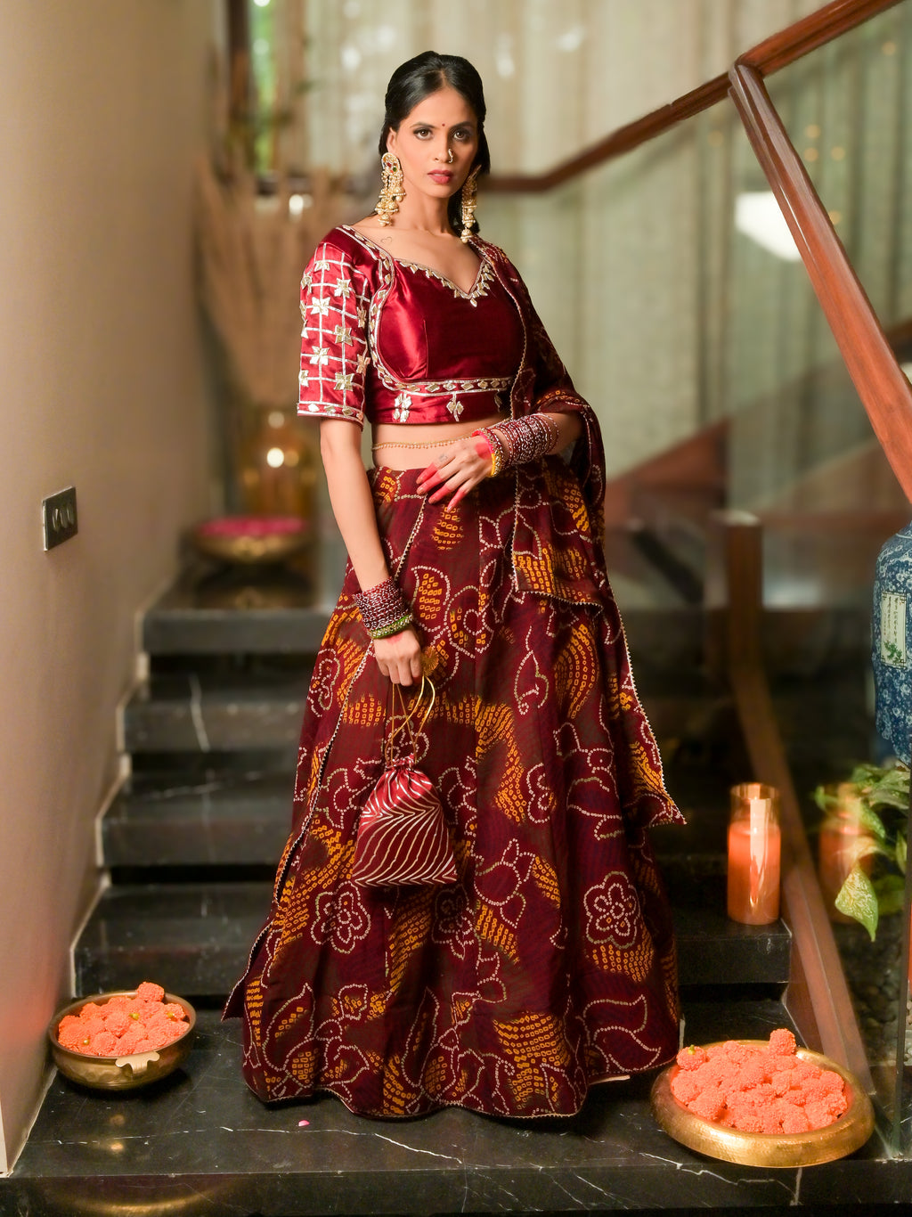 A woman stands on indoor stairs wearing a traditional Indian lehenga choli outfit in deep maroon and gold with floral patterns. The set includes a patterned blouse, a matching flared skirt, and a draped dupatta.
