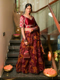 A woman stands on indoor stairs wearing a traditional Indian lehenga choli outfit in deep maroon and gold with floral patterns. The set includes a patterned blouse, a matching flared skirt, and a draped dupatta.