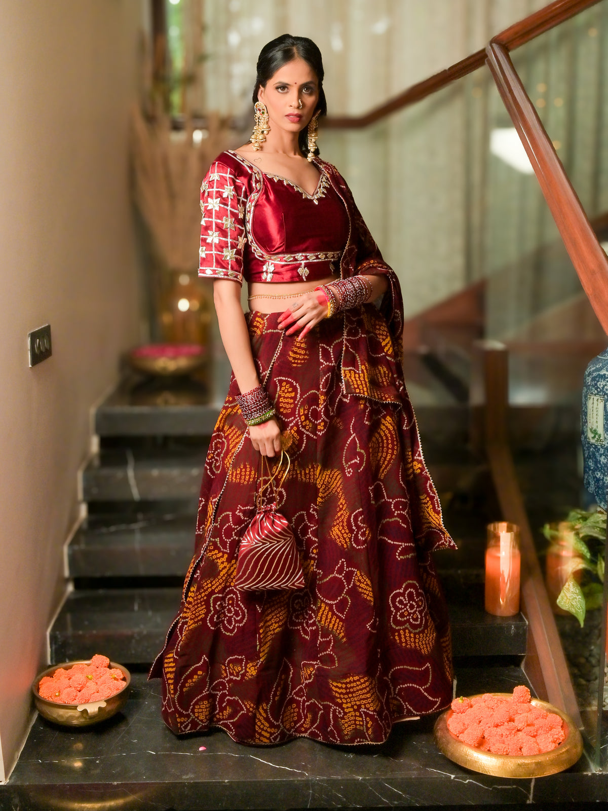 A woman stands on indoor stairs wearing a traditional Indian lehenga choli outfit in deep maroon and gold with floral patterns. The set includes a patterned blouse, a matching flared skirt, and a draped dupatta.