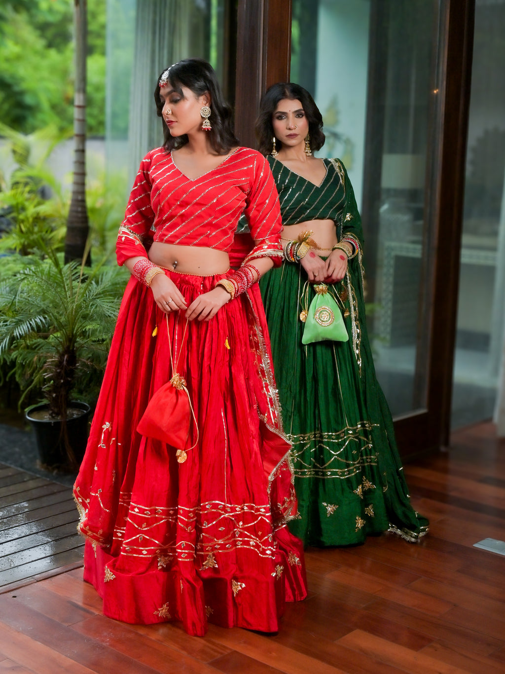 Two women modeling traditional Indian lehenga choli gowns in an indoor setting with a wooden floor and glass doors. The woman in the foreground wears a red gown with gold embroidery and holds a matching red handbag.
