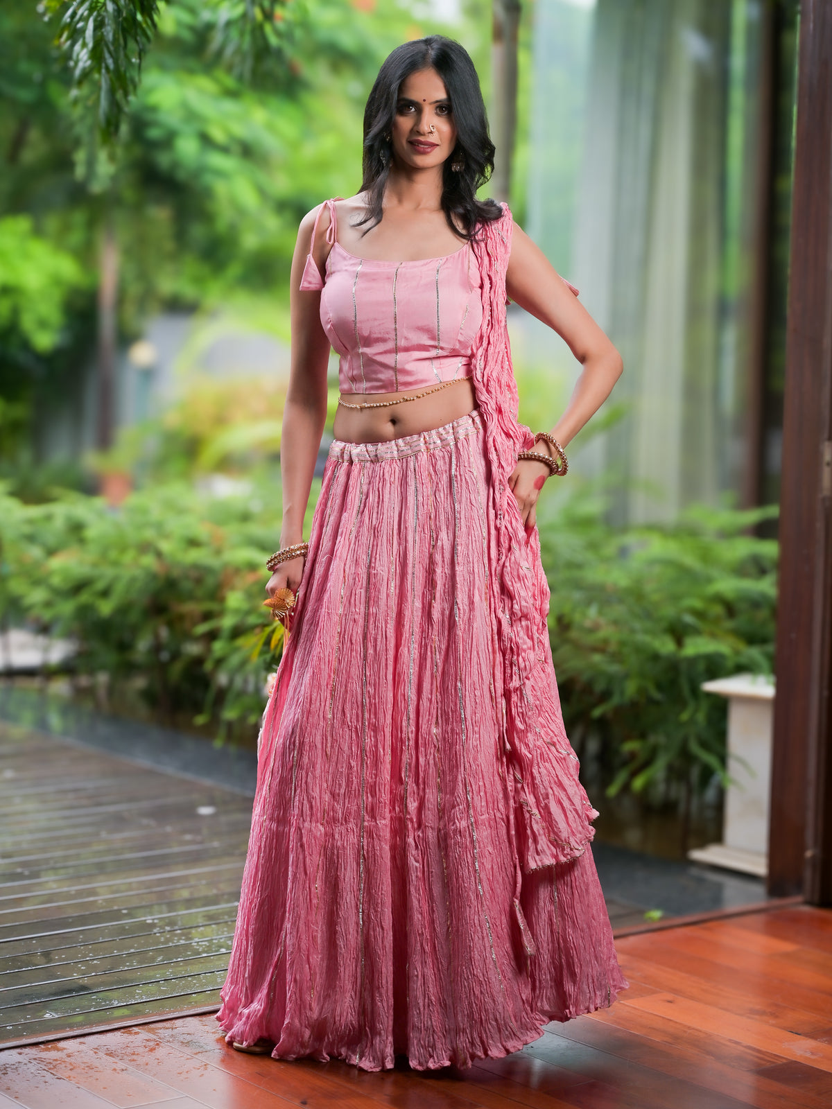 A woman modeling a pink traditional Indian outfit consisting of a sleeveless crop top, a long pleated skirt, and a draped dupatta, set against a lush green garden background.