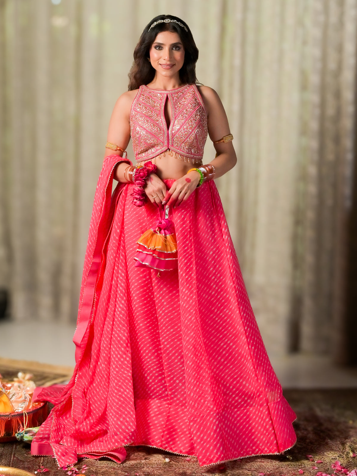 A woman modeling a bright pink traditional Indian lehenga choli outfit with intricate gold embroidery, a matching dupatta, and a small tassel accessory, set against a softly blurred indoor backdrop.