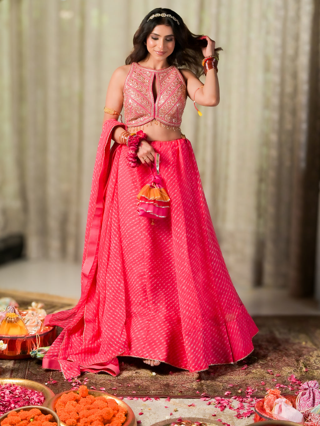 A woman models a vibrant pink traditional Indian lehenga choli outfit with intricate gold embroidery, standing in a festive setting decorated with flowers and ceremonial items.