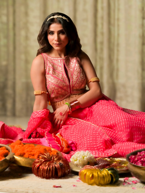 A woman wearing a vibrant pink and gold traditional Indian outfit sits on a floor surrounded by decorative pumpkins and bowls of flowers, set against a neutral draped background.