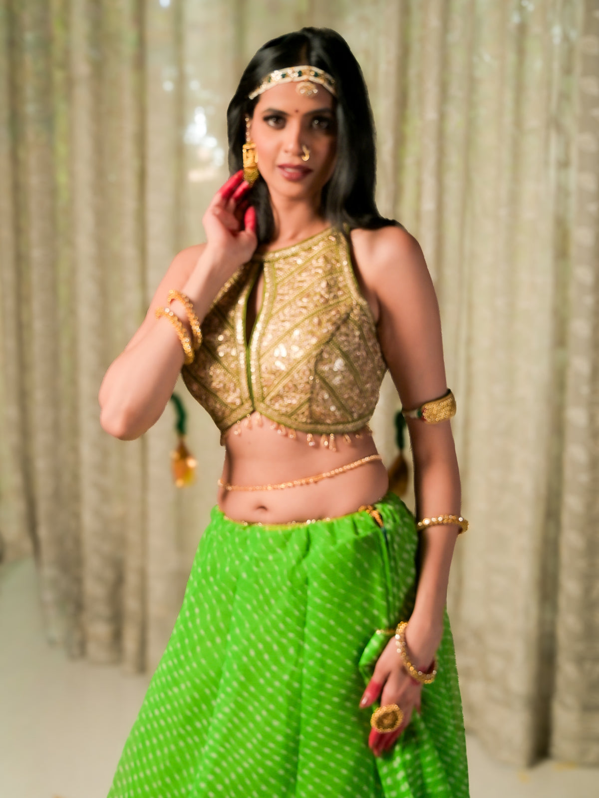 A woman modeling a traditional Indian outfit featuring a gold embellished crop top and a bright green skirt with white polka dots, accessorized with gold jewelry against a beige curtain background.