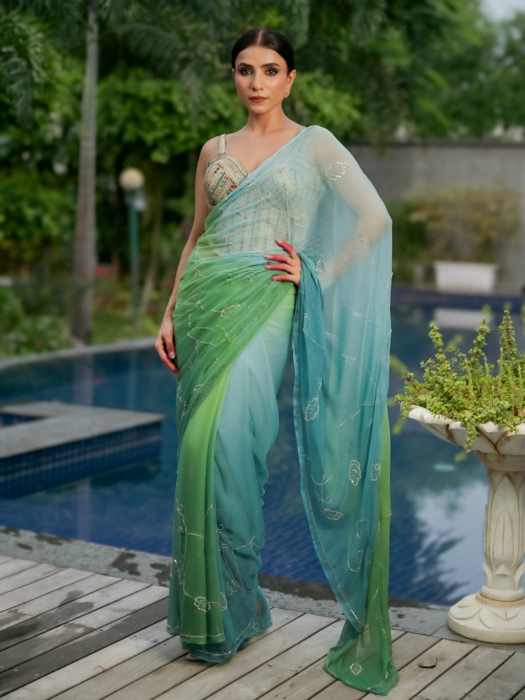 A woman models a sheer green and teal ombre saree with delicate gold embroidery, standing on a wooden deck next to a swimming pool with lush green garden foliage in the background.