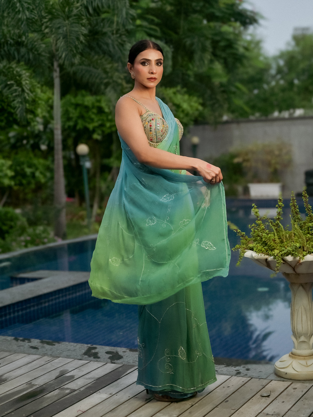 A woman stands by a swimming pool wearing a flowing green saree with a gradient from light aqua to deep green, featuring subtle embroidered patterns and a heavily embellished gold blouse.