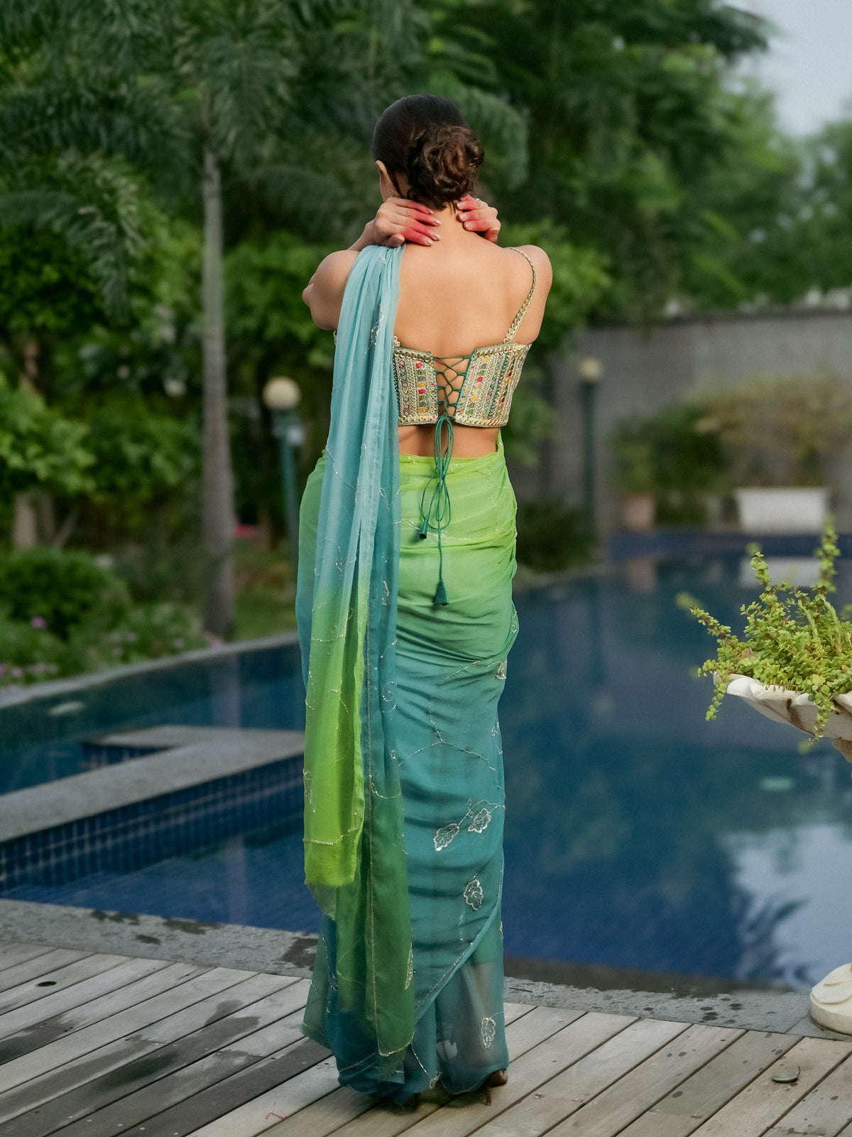 A woman stands with her back to the camera on a wooden deck beside a swimming pool, wearing a green and teal gradient saree with a patterned lace-up blouse.
