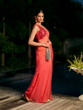A woman stands on a wooden deck beside a pool at night, wearing a coral-orange saree with a heavily embellished red and black sequined sleeveless blouse. She holds a silver patterned clutch.