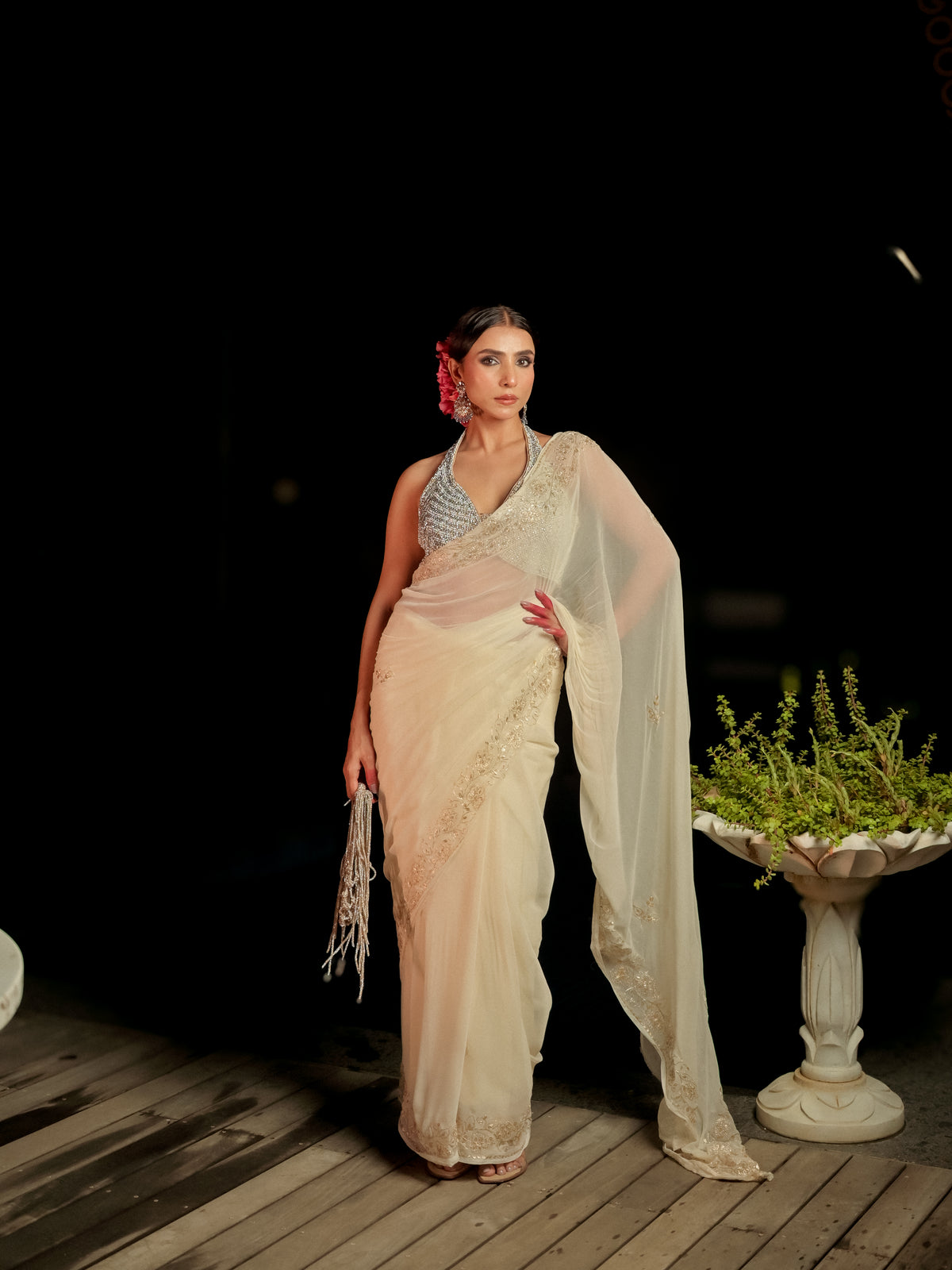 A woman models a traditional Indian saree with a light beige body, silver embellished border, and a sheer white dupatta, standing on a wooden deck against a dark background.