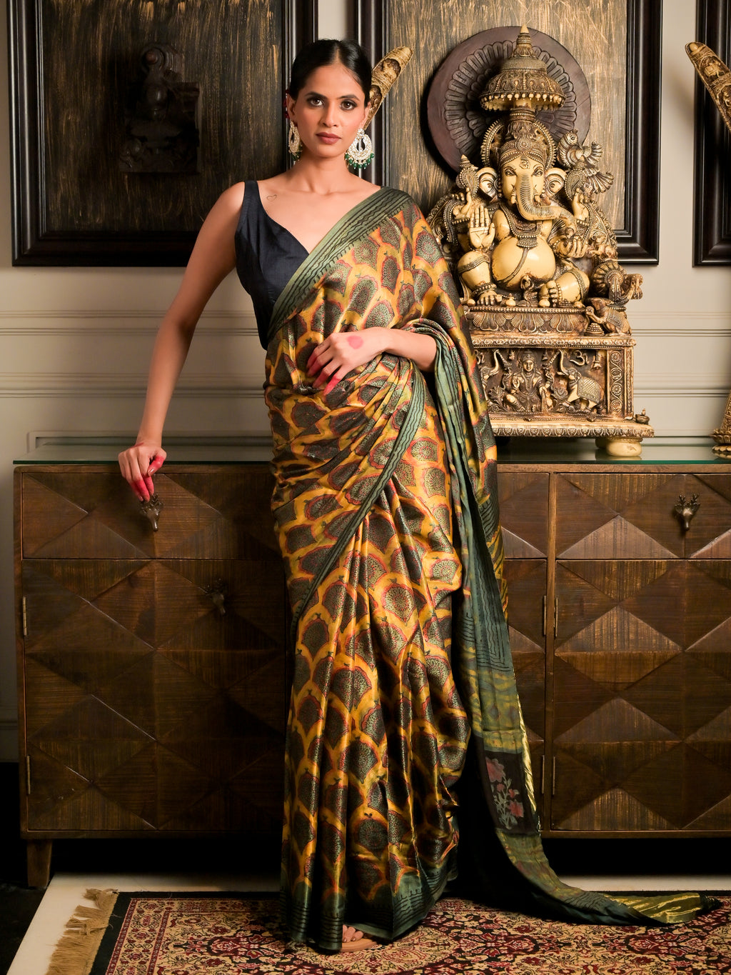 A woman models a dark green and gold patterned silk saree with a black sleeveless blouse, standing in front of a wooden cabinet with a Ganesha statue in a traditional Indian room.