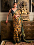 A woman models a dark green and gold patterned silk saree with a black sleeveless blouse, standing in front of a wooden cabinet with a Ganesha statue in a traditional Indian room.