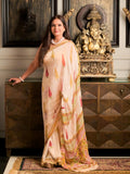 A woman wearing a light beige saree with pink and gold printed motifs stands indoors in front of a dark wooden cabinet and a gold Ganesha statue.