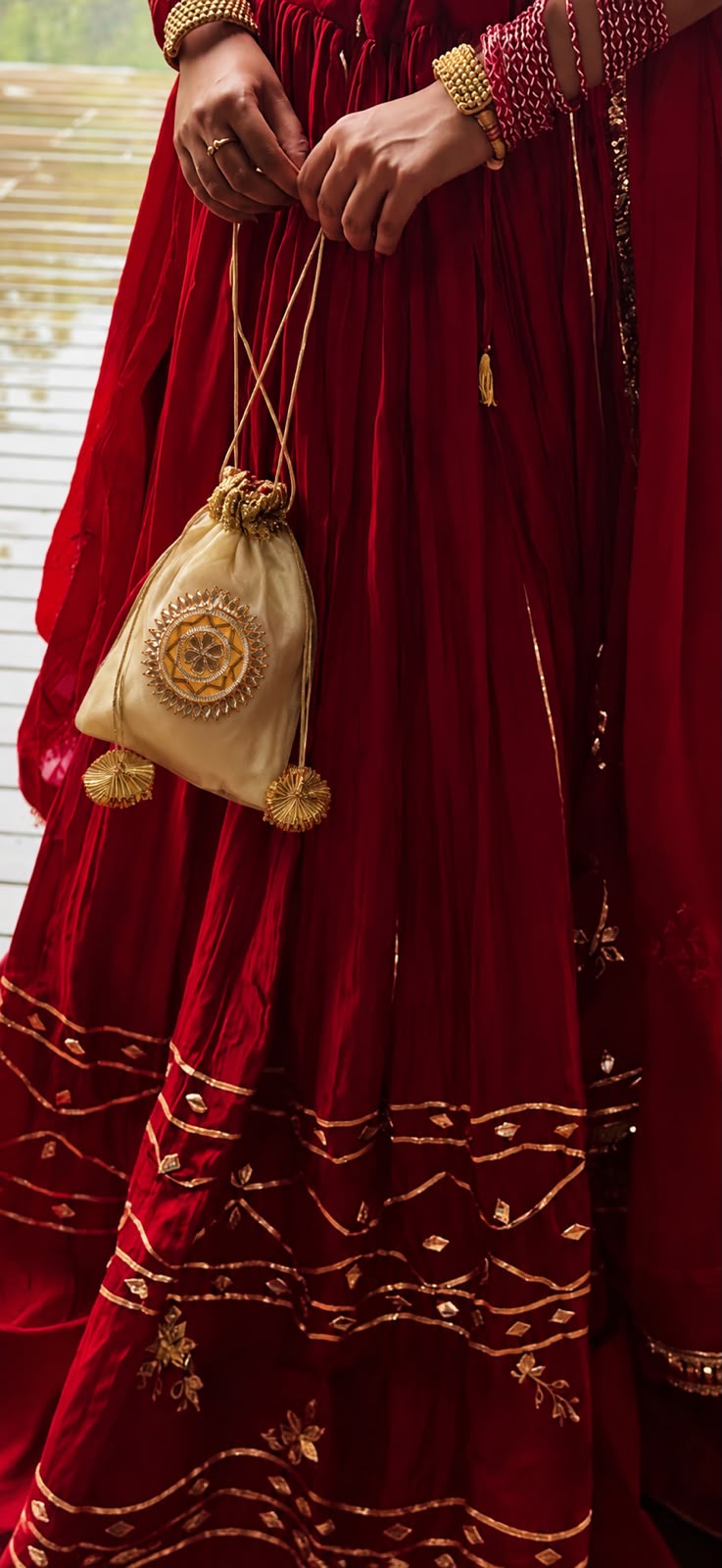 A close-up lifestyle shot of a person wearing a traditional deep red embroidered dress and holding a small beige drawstring pouch decorated with gold embroidery and tassels.