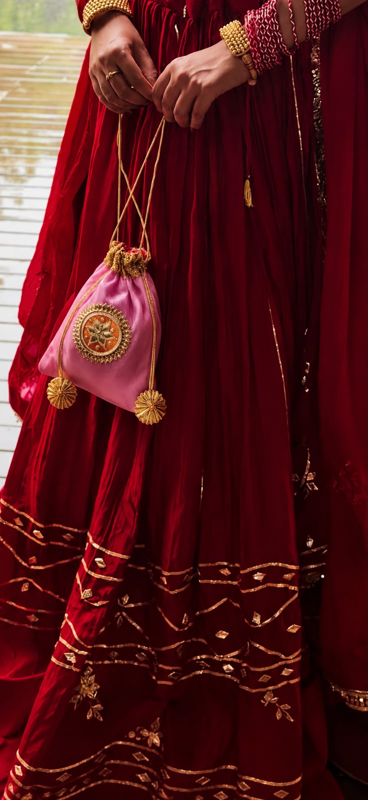 A close-up lifestyle shot of a person wearing a traditional deep red embroidered dress and holding a small pink drawstring pouch decorated with gold embroidery and tassels.