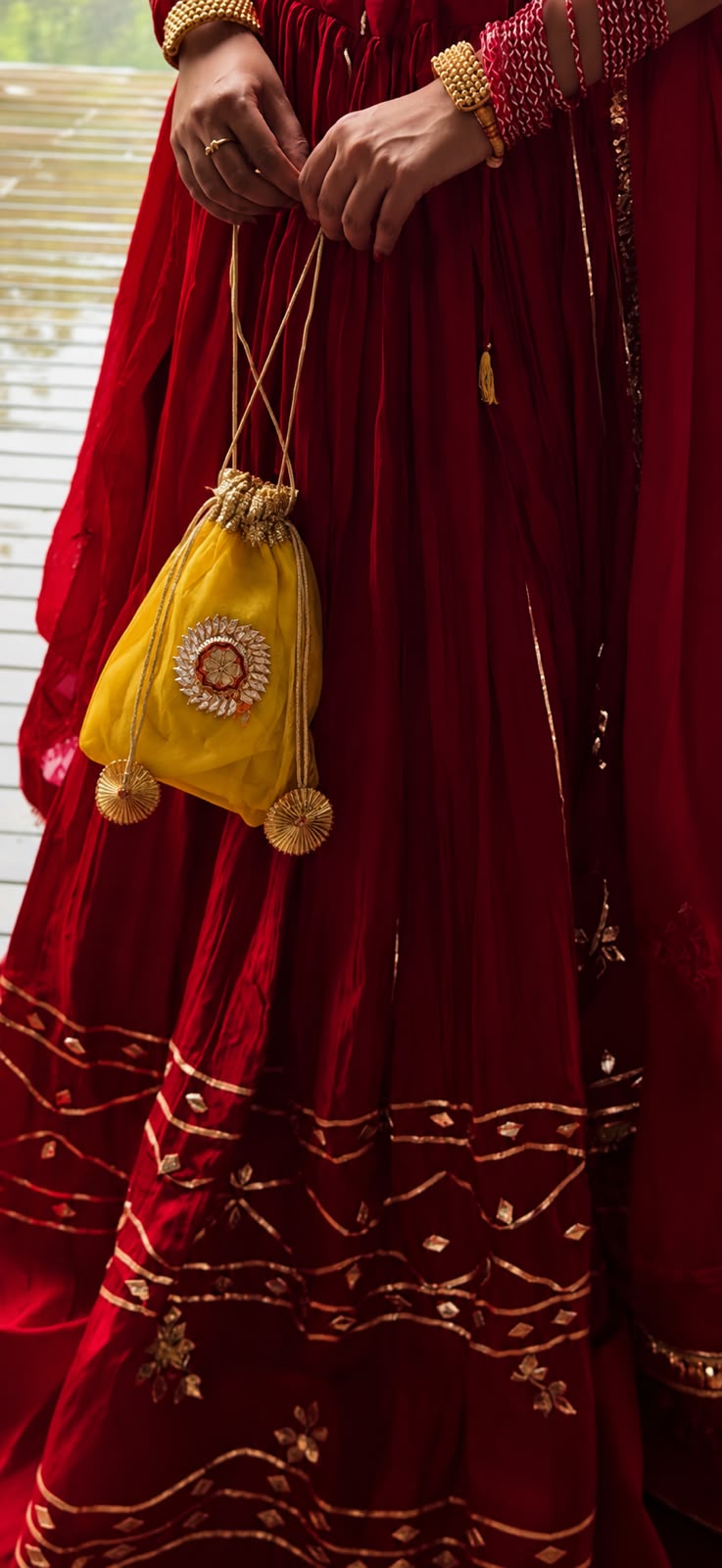 A close-up lifestyle shot of a person wearing a traditional deep red embroidered skirt and holding a small yellow drawstring pouch decorated with gold embroidery and tassels.