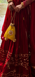 A close-up lifestyle shot of a person wearing a traditional deep red embroidered skirt and holding a small yellow drawstring pouch decorated with gold embroidery and tassels.