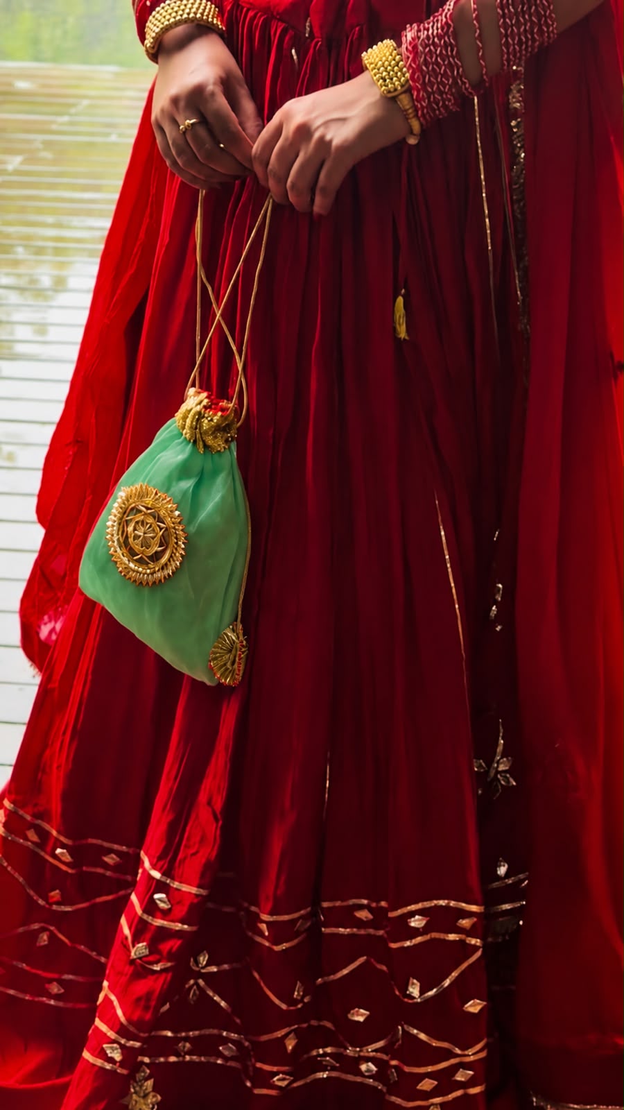 A close-up of a person wearing a rich red traditional dress with gold embroidery, holding a small mint green drawstring pouch decorated with gold hardware.
