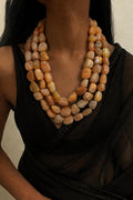 A close-up of a model wearing a multi-strand necklace made of polished, peachy-orange and beige stone beads, paired with a black sheer top.